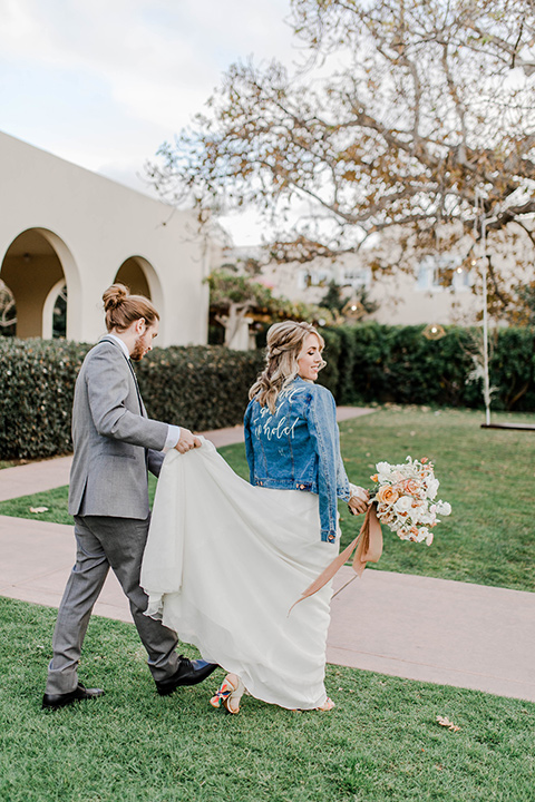 bride and groom walking, the bride in a white gown with an illusion neckline and a jean jacket and the groom in a grey suit with black trim and a black long tie
