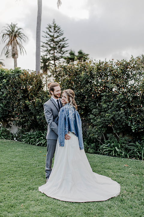 bride and groom holding each other, the bride in a white lace gown with a jean jacket and the groom in a grey suit with black trim and black long tie