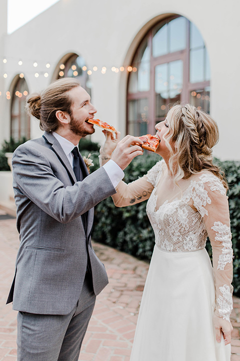 bride in a white lace gown with an illusion neckline and the groom in a grey suit with a black trim and black long tie eating pizza