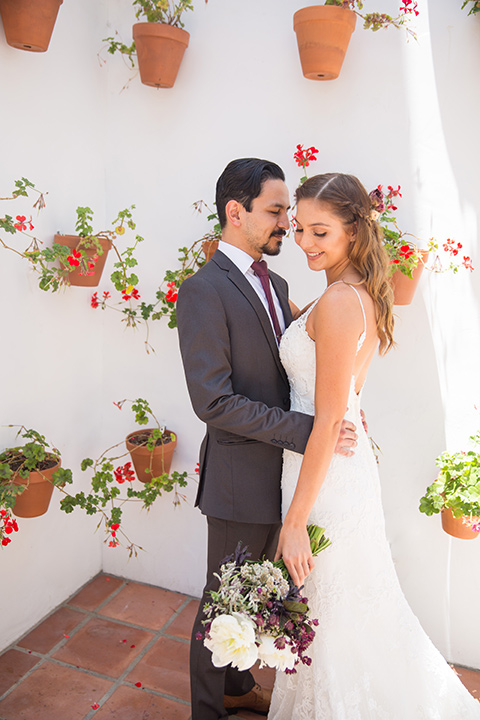 bride in a white lace gown with thin straps and a white and red floral bouquet and the groom in a grey suit with a red long tie