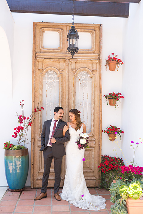 bride in a white lace gown with thin straps and a white and red floral bouquet and the groom in a grey suit with a red long tie by wooden door