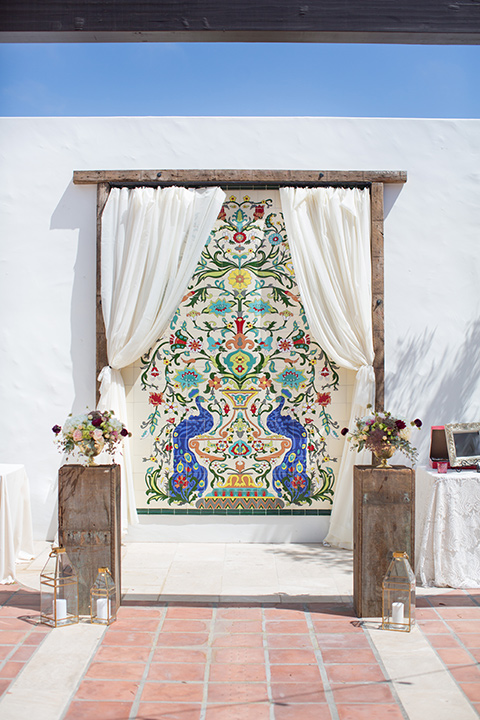 ceremony arch with wood and white flowing linens and colorful tile wall behind them