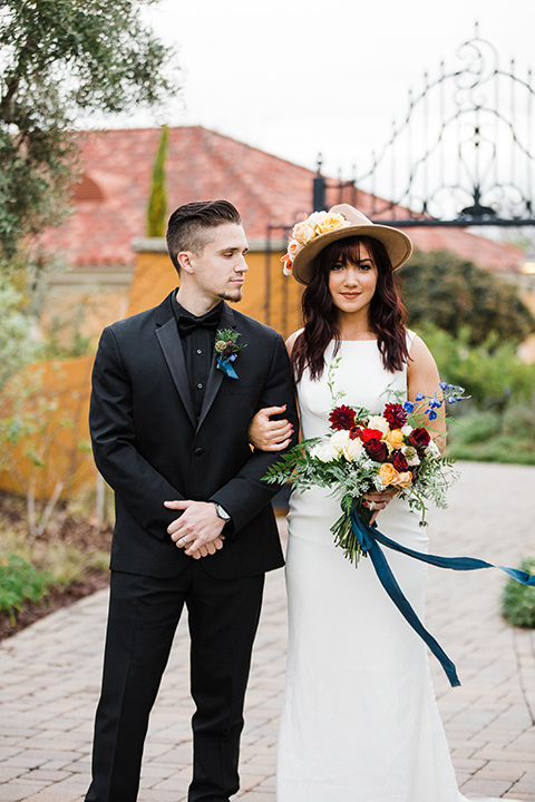 bride and groom standing facing camera bride in a bohemian style dress with a high neckline and lace with a hat groom in an all-black tuxedo look with a black shirt and bow