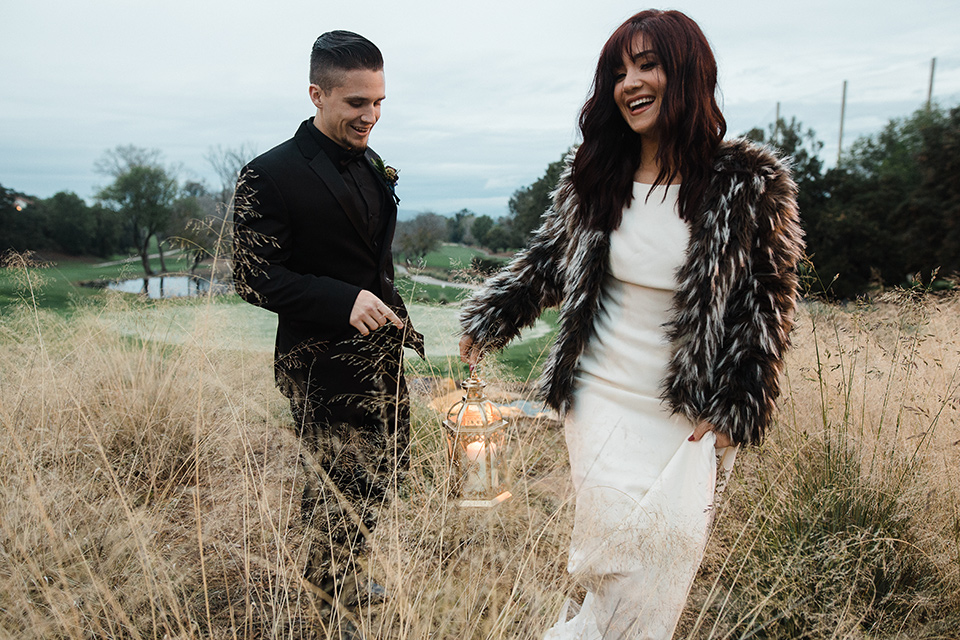 Los Robles Greens Shoot bride and groom walking in tall grass bride in a bohemian style gown with lace and a high neckline groom in a black tuxedo with a black shirt and bow tie