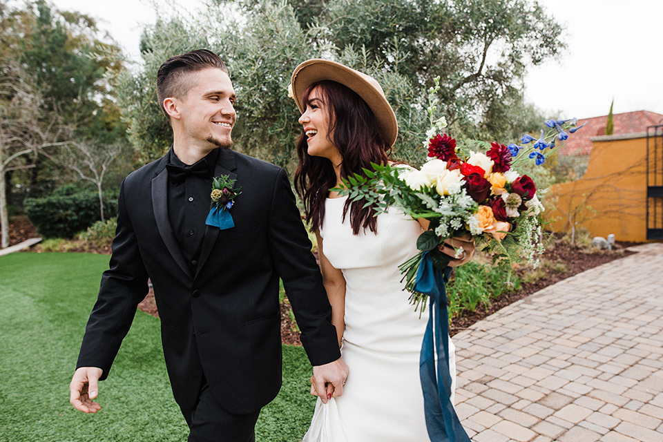 bride and groom walking bride in a bohemian style gown with lace and a high neckline groom in a black tuxedo with a black shirt and bow tie