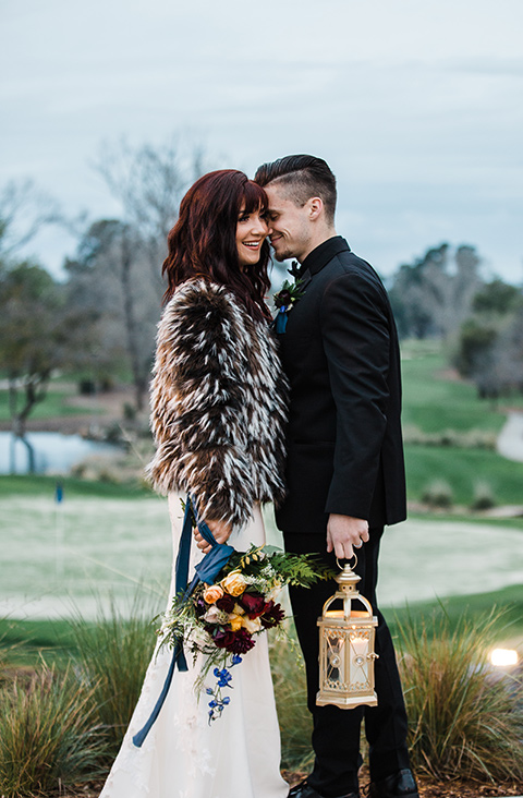 bride in fur holding lantern bride in a bohemian style dress with a high neckline and lace with a hat groom in an all black tuxedo look with a black shirt and bow