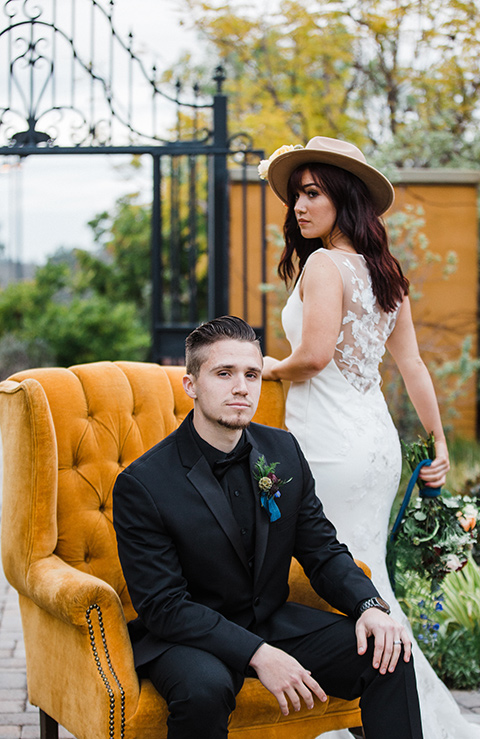 groom sitting bride stands bride in a bohemian style dress with a high neckline and lace with a hat groom in an all black tuxedo look with a black shirt and bow