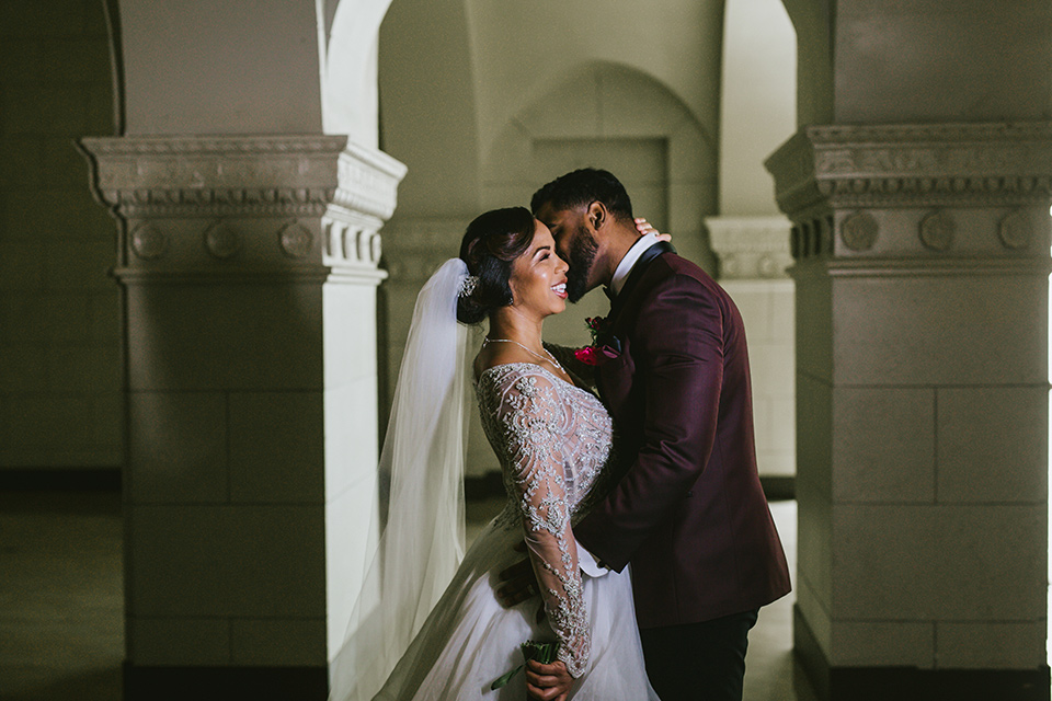 majestic-downtown-los-angeles-shoot-bride-and-groom-close-embrace-bride-in-a-tulle-ballgown-with-a-beaded-bodice-with-long-sleeves-groom-in-a-burgundy-tuxedo-with-a-shawl-lapel-and-black-bow-tie