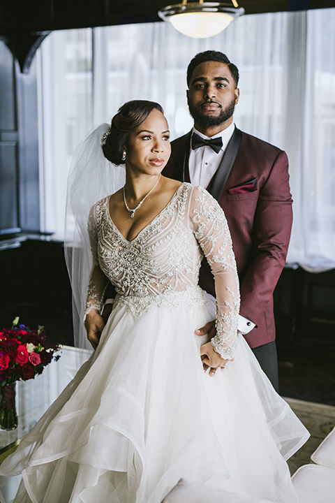 majestic-downtown-los-angeles-shoot-bride-and-groom-standing-groom-behind-bride-bride-in-a-tulle-ballgown-with-a-beaded-bodice-with-long-sleeves-and-the-groom-in-a-burgundy-tuxedo-with-a-black-satin-lapel-and-black-bow-tie