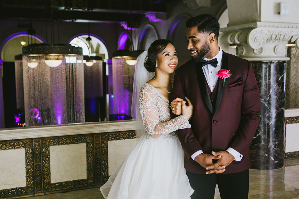 majestic-downtown-los-angeles-shoot-bride-behind-groom-groom-smiling-at-bride-bride-and-groom-close-embrace-bride-in-a-tulle-ballgown-with-a-beaded-bodice-with-long-sleeves-groo-in-a-burgundy-tuxedo-with-a-satin-lapel-and-black-bow-tie