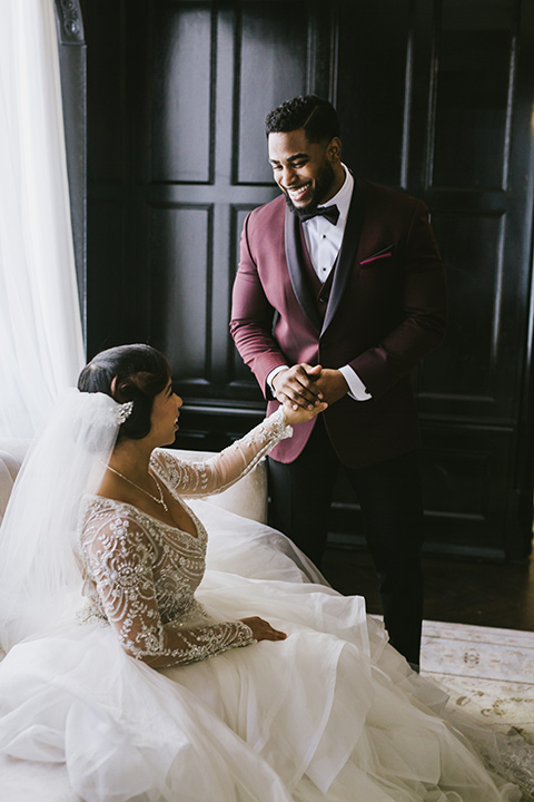 majestic-downtown-los-angeles-shoot-bride-sitting-groom-standing-looking-at-her-the-bride-in-a-tulle-ball-gown-with-beaded-bodice-and-long-sleeves-the-groom-in-a-burgundy-tuxedo-with-a-black-shawl-lapel-and-black-bow-tie