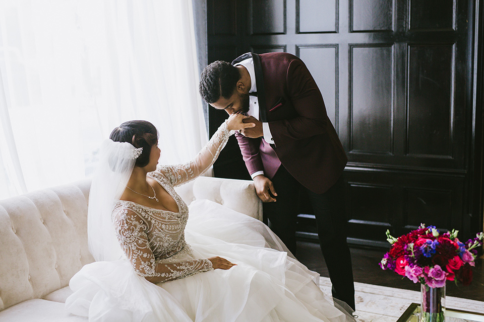 majestic-downtown-los-angeles-shoot-groom-kissing-brides-hand-bride-in-a-tulle-ballgown-with-a-beaded-bodice-with-long-sleeves-groo-in-a-burgundy-tuxedo-with-a-satin-lapel-and-black-bow-tie