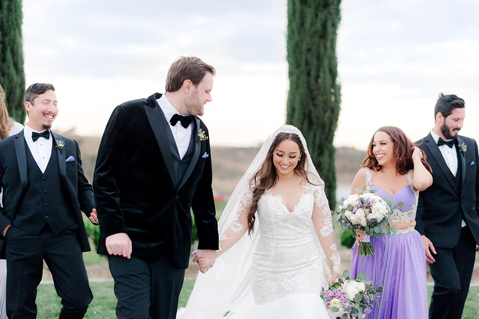  bride in a white lace gown with long sleeves and a tulle skirt and the groom in a black velvet tuxedo with a black bow tie, the bridesmaids in purple gowns and the groomsmen in black tuxedos 