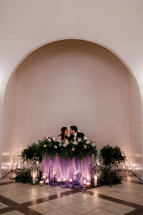  bride in a white lace gown with long sleeves and a tulle skirt and the groom in a black velvet tuxedo with a black bow tie 