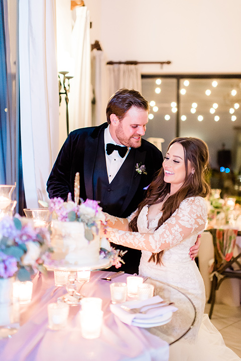  bride in a white lace gown with long sleeves and a tulle skirt and the groom in a black velvet tuxedo with a black bow tie cutting a cake