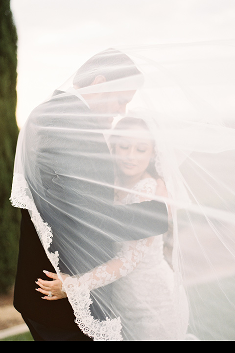  bride in a white lace gown with long sleeves and a tulle skirt and the groom in a black velvet tuxedo with a black bow tie 