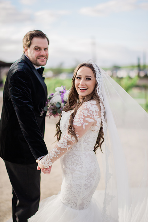  bride in a white lace gown with long sleeves and a tulle skirt and the groom in a black velvet tuxedo with a black bow tie cutting a cake