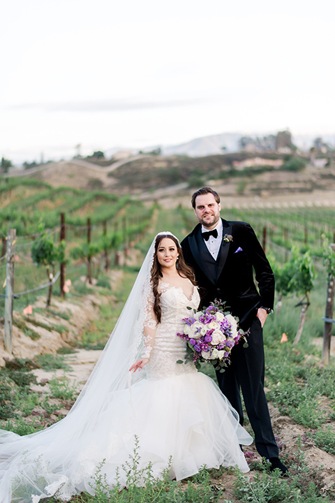  bride in a white lace gown with long sleeves and a tulle skirt and the groom in a black velvet tuxedo with a black bow tie 