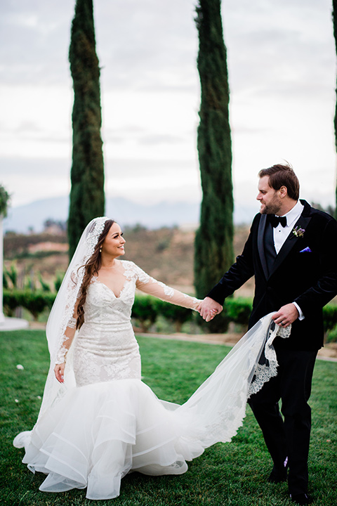  bride in a white lace gown with long sleeves and a tulle skirt and the groom in a black velvet tuxedo with a black bow tie cutting a cake