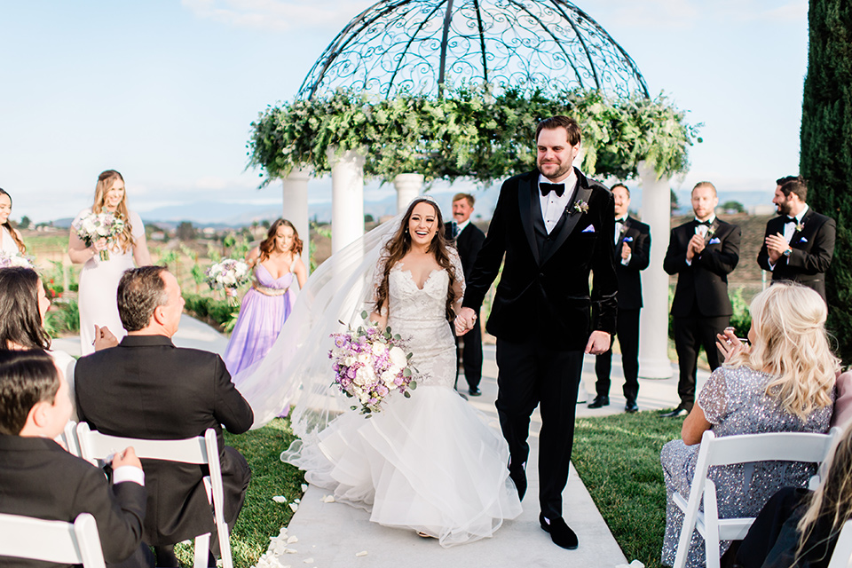  bride in a white lace gown with long sleeves and a tulle skirt and the groom in a black velvet tuxedo with a black bow tie, the bridesmaids in purple gowns and the groomsmen in black tuxedos 