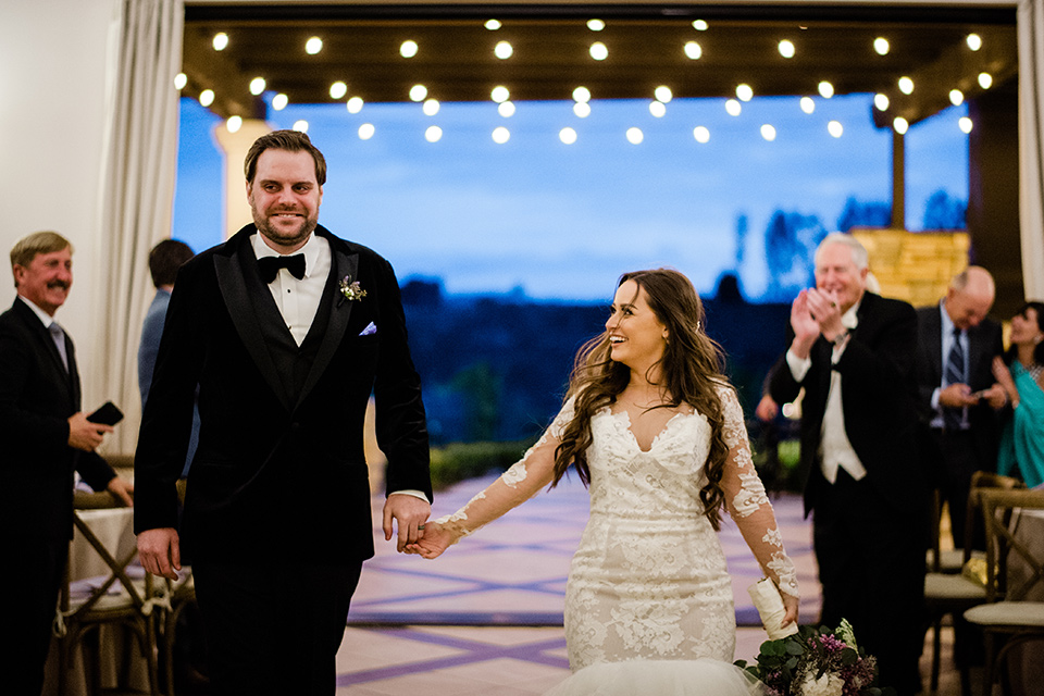  bride in a white lace gown with long sleeves and a tulle skirt and the groom in a black velvet tuxedo with a black bow tie, the bridesmaids in purple gowns and the groomsmen in black tuxedos 