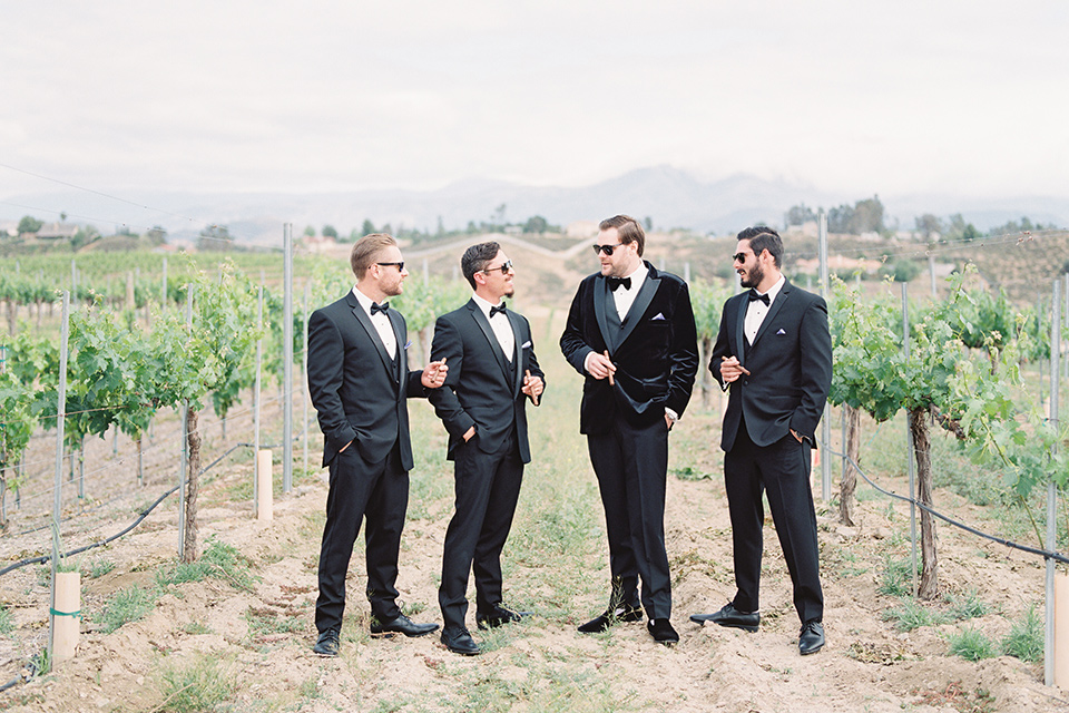  the groom in a black velvet tuxedo with a black bow tie, the and the groomsmen in black tuxedos 