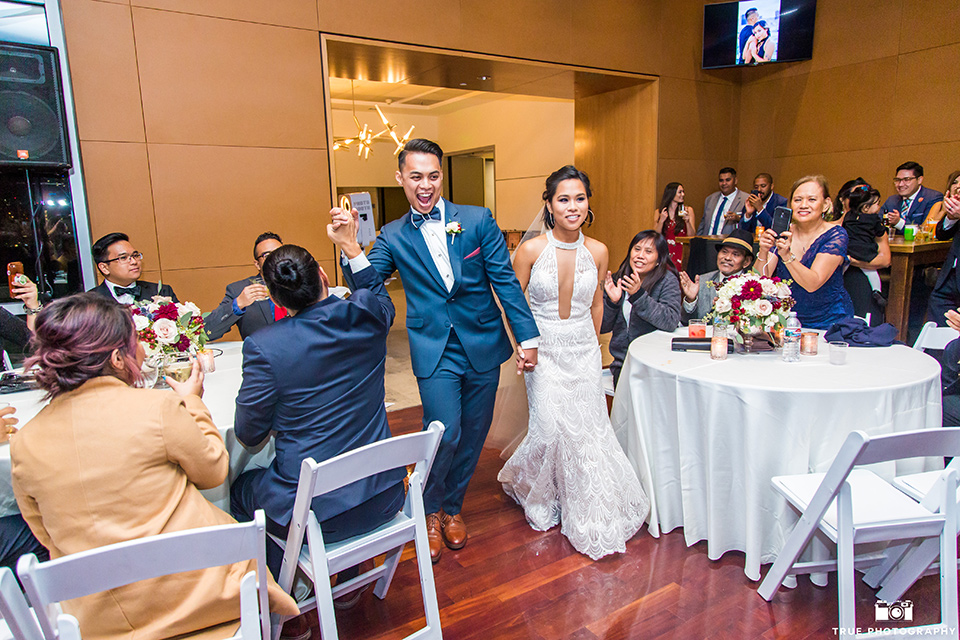 skybox-real-wedding-bride-and-groom-enter-reception-bride-wearing-a-fitting-lace-dress-with=straps-groom-wearing-a-blue-suit-bridesmaids-in-burgundy-groomsmen-in-blue