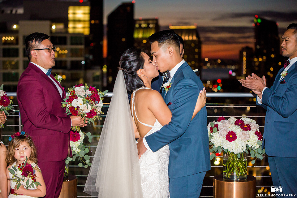 skybox-real-wedding-bride-and-groom-first-kiss-bride-wearing-a-fitting-lace-dress-with=straps-groom-wearing-a-blue-suit-bridesmaids-in-burgundy-groomsmen-in-blue