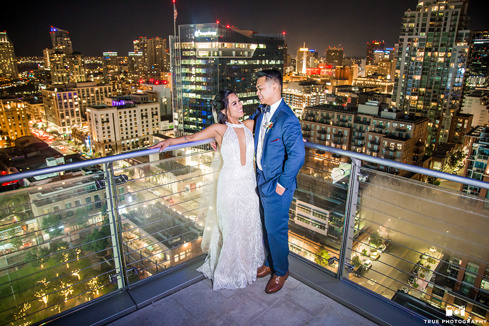 skybox-real-wedding-bride-and-groom-with-lighted-up-city-behind-them-bride-wearing-a-fitting-lace-dress-with=straps-groom-wearing-a-blue-suit-bridesmaids-in-burgundy-groomsmen-in-blue