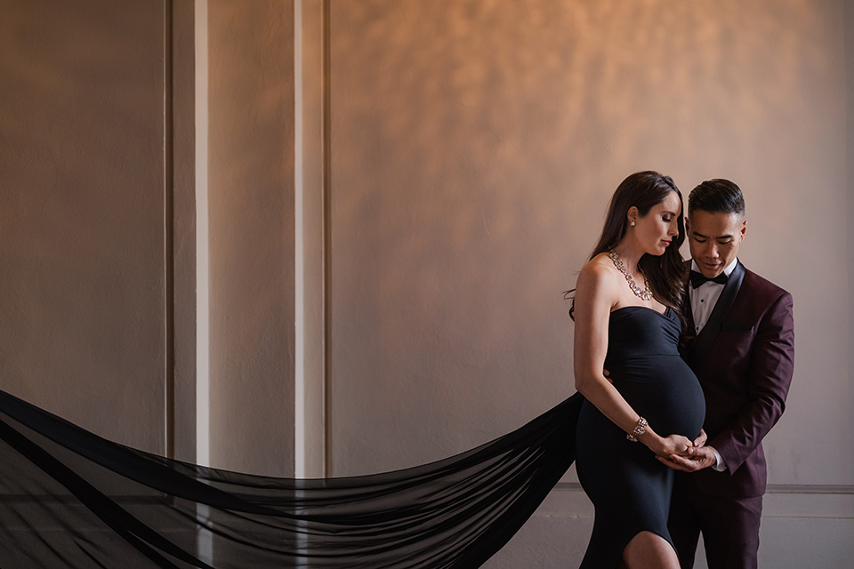 Maternity-workshop-husband-and-wife-holding-eachother-her-gown-flowing-mom-wearing-a-chic-black-formfittng-gown-dad-wearing-a-burgundy-tuxedo-and-black-bow-tie