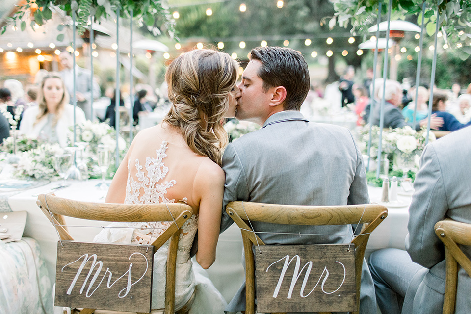 Temecula-Creek-Inn-Wedding-bride-and-groom-kissing-at-sweetheart-table-bride-in-a-form-fitting-lace-gown-with-an-illusion-back-detail-groom-in-a-light-grey-suit-with-a-light-blue-polka-dot-tie