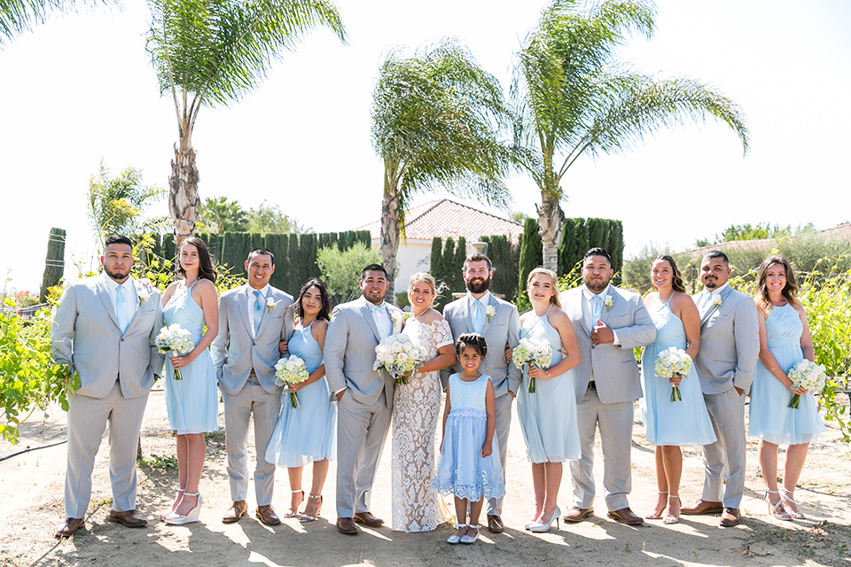 bride in a lace formfitting gown with an illusion neckline the groom in a light grey suit and bow tie, the groomsmen in light grey suits and blue ties and the brides maids in light blue short gowns 