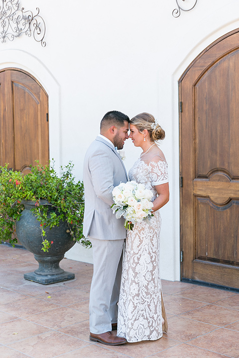  bride in a lace formfitting gown with an illusion neckline the groom in a light grey suit and bow tie 