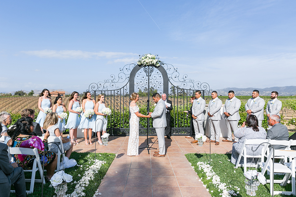 bride in a lace formfitting gown with an illusion neckline the bridesmaids in light blue short gowns groomsmen in light grey suits and groom in a light grey suit 