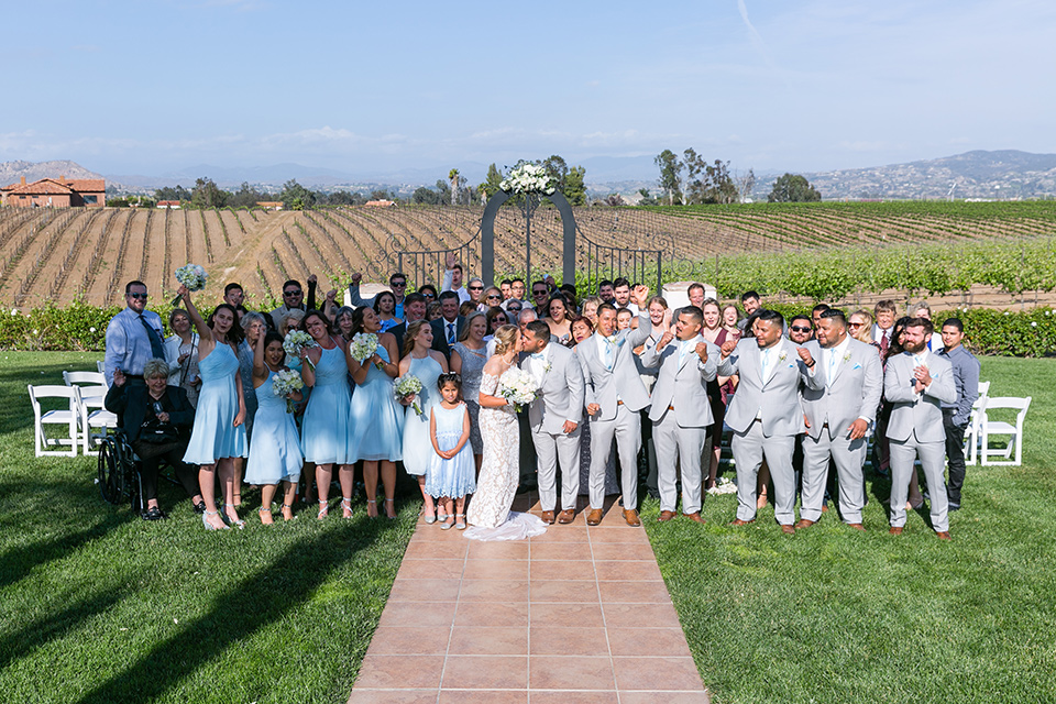  groom and groomsmen in a light grey suit and bow tie and light blue long ties bridesmaids in light blue suits and all the guests smiling