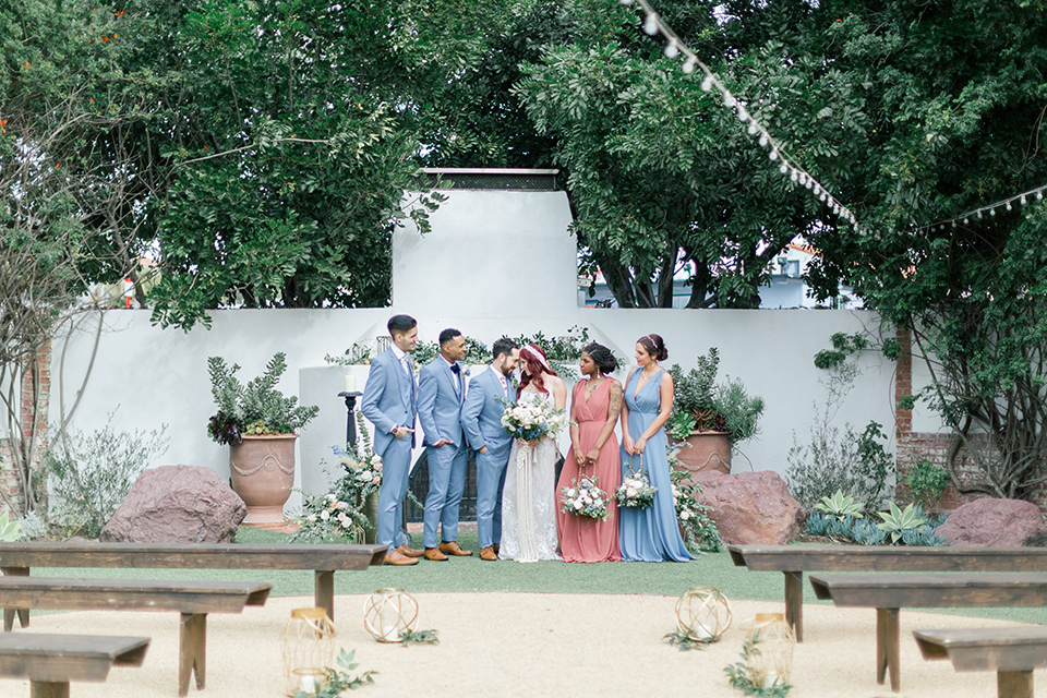  bride in a cream form fitting lace gown with a macramé headpiece, groom in a light blue suit with a floral tie, groomsmen in light blue suits and navy bow ties, bridesmaids in light blue and coral gowns 