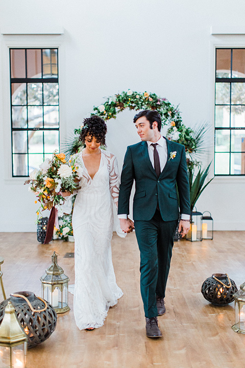 modern Moroccan shoot bride and groom at ceremony space walking down the aisle the bride in a white lace gown with long sleeves and a v neck line and the groom is in a green suit with a brown long tie