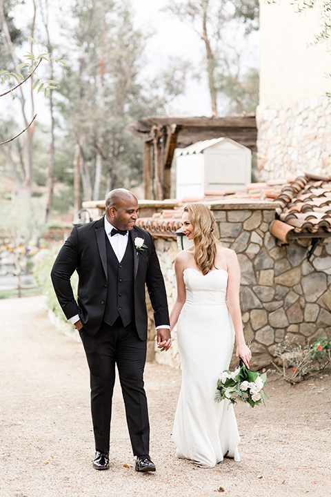 Oak-Meadows-Shoot-bride-and-groom-walking-by-venue-bride-wearing-a-lace-strapless-dress-groom-in-a-black-notch-lapel-tuxedo-with-a-black-bow-tie