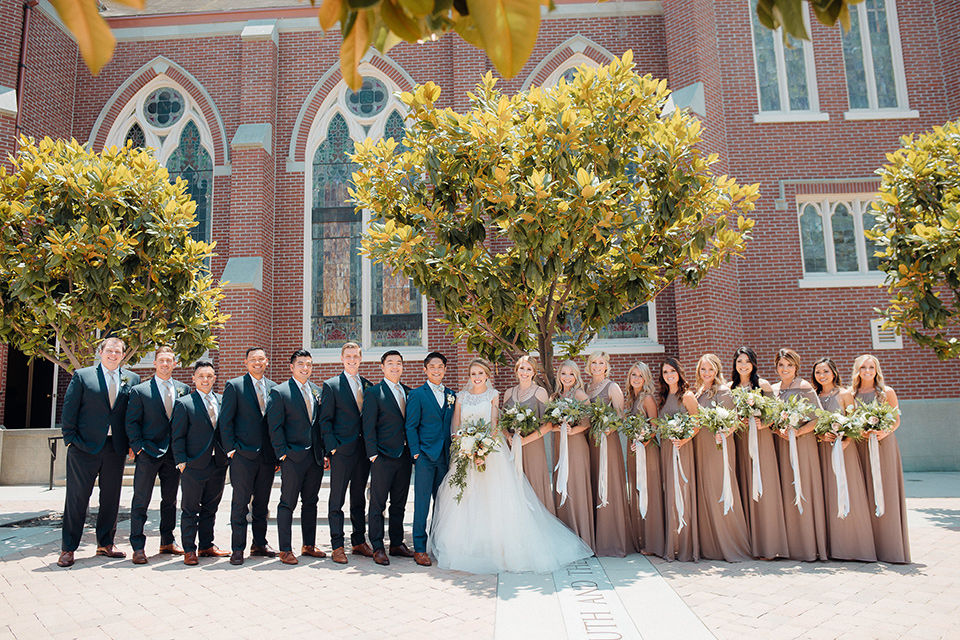 orange-county-wedding-bridal-party-outside-church-bridesmaids-in-a-taupe-dress-color-bride-in-a-two-piece-lace-ballgown-groomsmen-in-a-dark-navy-suit-groom-in-a-brighter-blue-suit-with-a-flowal-bowtie