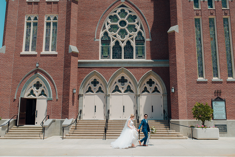 orange-county-wedding-bride-and-groom-walking-in-front-of-church-bride-in-a-two-piece-lace-ballgown-groom-in-a-brighter-blue-suit-with-a-flowal-bowtie