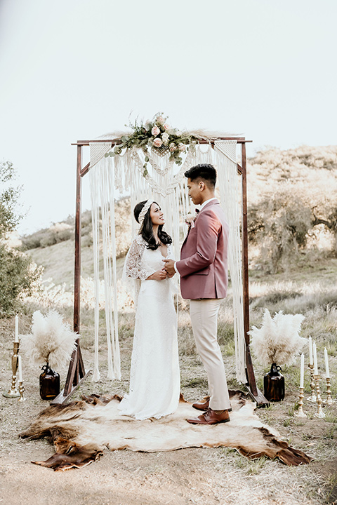 Owl-Creek-Farms-shoot-bride-and-groom-at-ceremony-bride-wearing-a-bohemian-style-dress-with-flutter-sleeves-and-a-macrame-style-headpiece-groom-in-a-rose-pink-suit-jacket-with-tan-pants-and-a-velvet-bow-tie