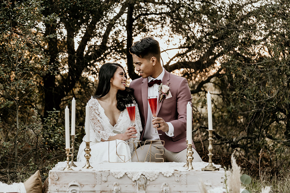 Owl-Creek-Farms-shoot-bride-and-groom-at-sweetheart-table-bride-wearing-a-bohemian-style-dress-with-flutter-sleeves-and-a-macrame-style-headpiece-groom-in-a-rose-pink-suit-jacket-with-tan-pants-and-a-velvet-bow-tie