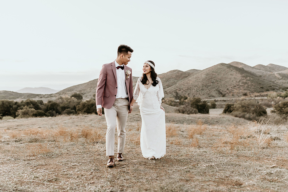 Owl-Creek-Farms-shoot-bride-and-groom-walking-bride-wearing-a-bohemian-style-dress-with-flutter-sleeves-and-a-macrame-style-headpiece-groom-in-a-rose-pink-suit-jacket-with-tan-pants-and-a-velvet-bow-tie
