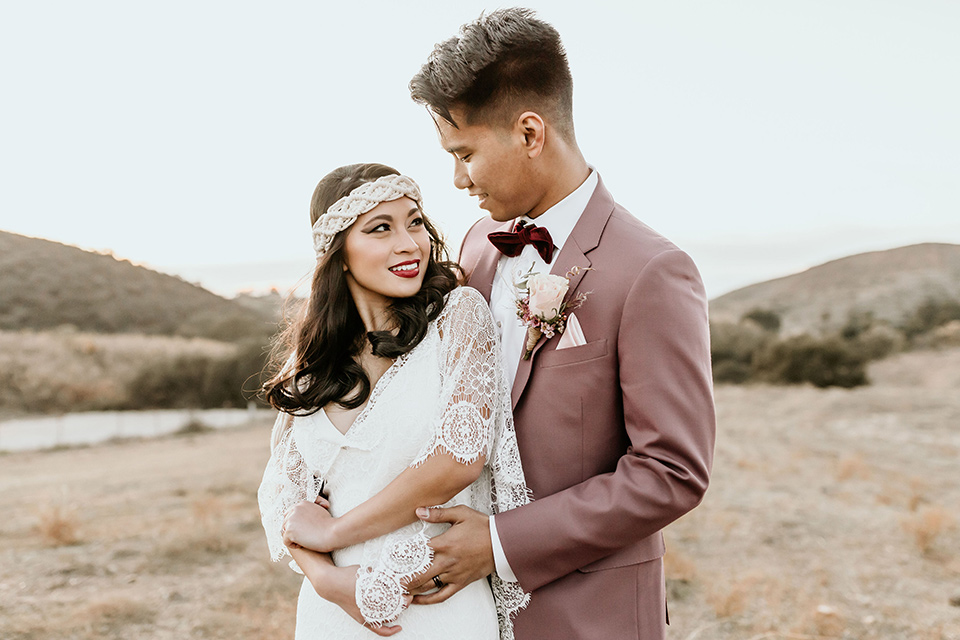 Owl-Creek-Farms-shoot-groom-holding-bride-while-the-bride-looks-at-her-bride-wearing-a-bohemian-style-dress-with-flutter-sleeves-and-a-macrame-style-headpiece-groom-in-a-rose-pink-suit-jacket-with-tan-pants-and-a-velvet-bow-tie