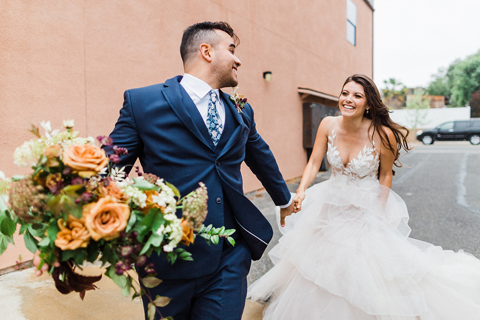 groom in a deep blue suit and floral tie running with bride in a white tulle ballgown