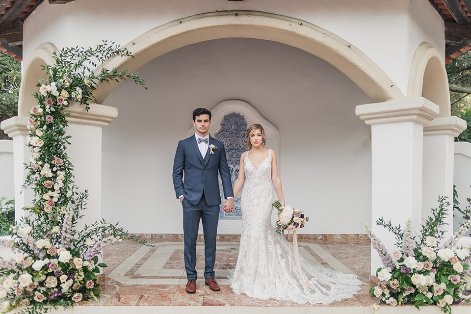 bride-and-groom-standing-facing-camera-while-the-bride-is-wearing-a-lace-dress-with-straps-and-illusion-back-groom-in-a-dark-blue-suit-with-a-grey-velvet-bow-tie