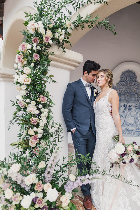 rancho-las-lomas-wedding-bride-and-groom-by-floral-arrangement-bride-wearing-a-dress-with-an-illusion-detail-and-straps-with-lace-on-the-dress-groom-wearing-a-dark-blue-suit-with-a-grey-velvet-bow-tie