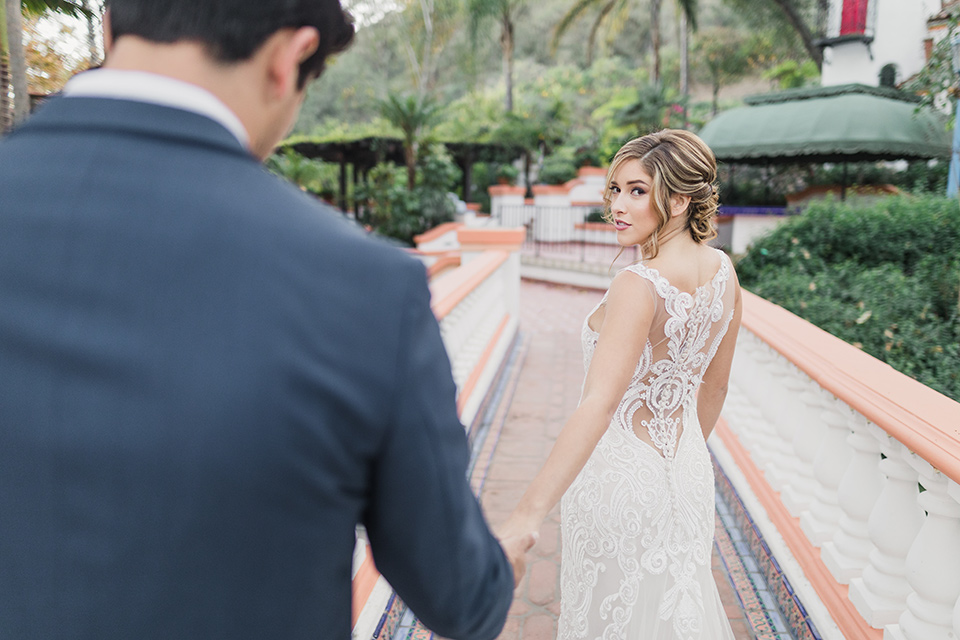 rancho-las-lomas-wedding-bride-taking-grooms-hand-bride-is-wearing-a-lace-dress-with-straps-and-illusion-back-groom-in-a-dark-blue-suit-with-a-grey-velvet-bow-tie