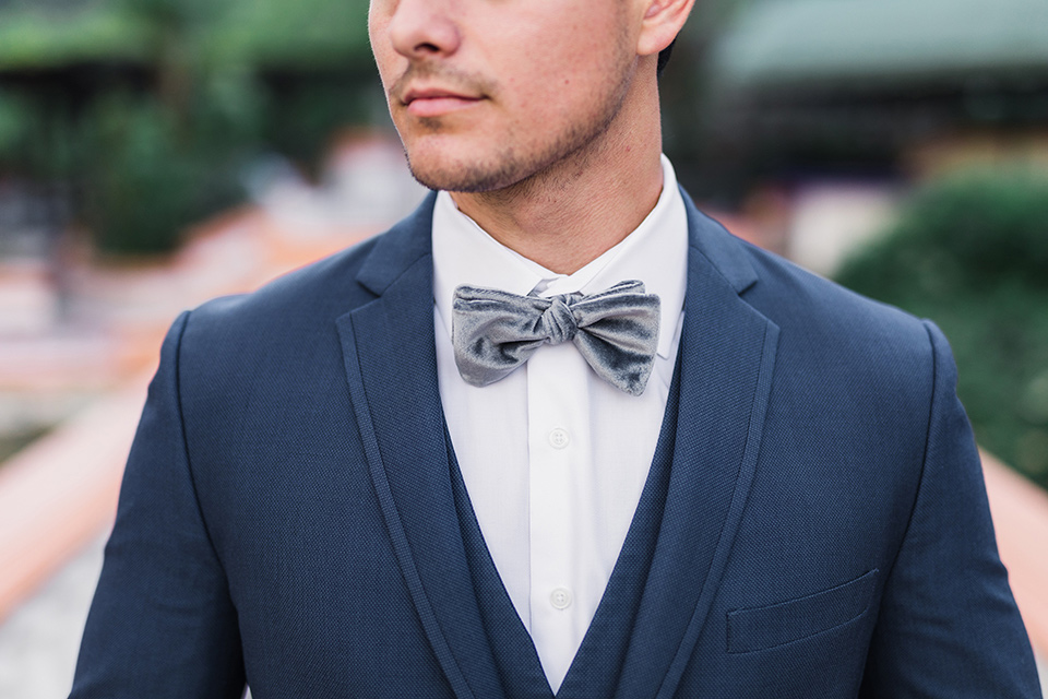 rancho-las-lomas-wedding-close-up-on-groom-attire-groom-in-a-dark-blue-suit-with-a-grey-velvet-bow-tie