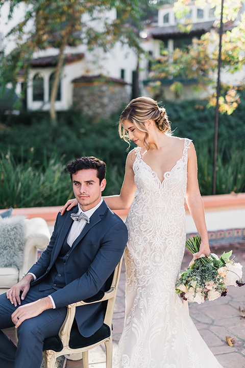 rancho-las-lomas-wedding-groom-sitting-bride-standing-looking-down-at-him-bride-in-lace-with-dress-groom-in-blue-suit-with-grey-bowtie